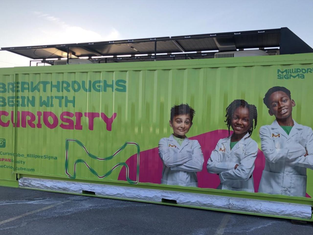 A green shipping container turned mobile science lab features three young scientists and text reading, "Breakthroughs Begin With Curiosity."