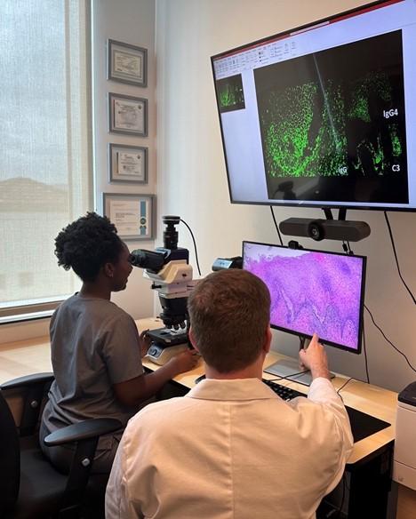 A person looks through a microscope while a person in a white coat points to a screen
