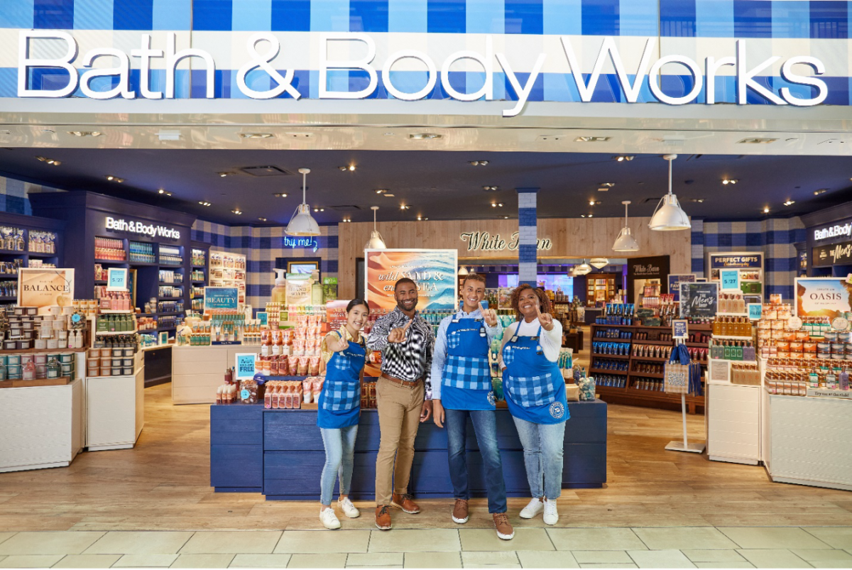 Four Bath & Body Works associates stand at the entrance of a store smiling and giving a “number 1” hand signal to the camera. A colorful selection of candles, hand soaps, lotions and other products are displayed on tables behind them.