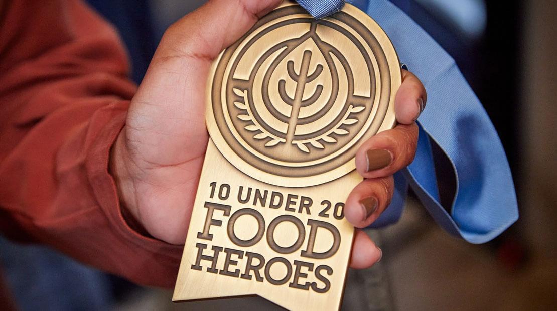 A hand holding a medal "10 Under 20 Food Heroes".