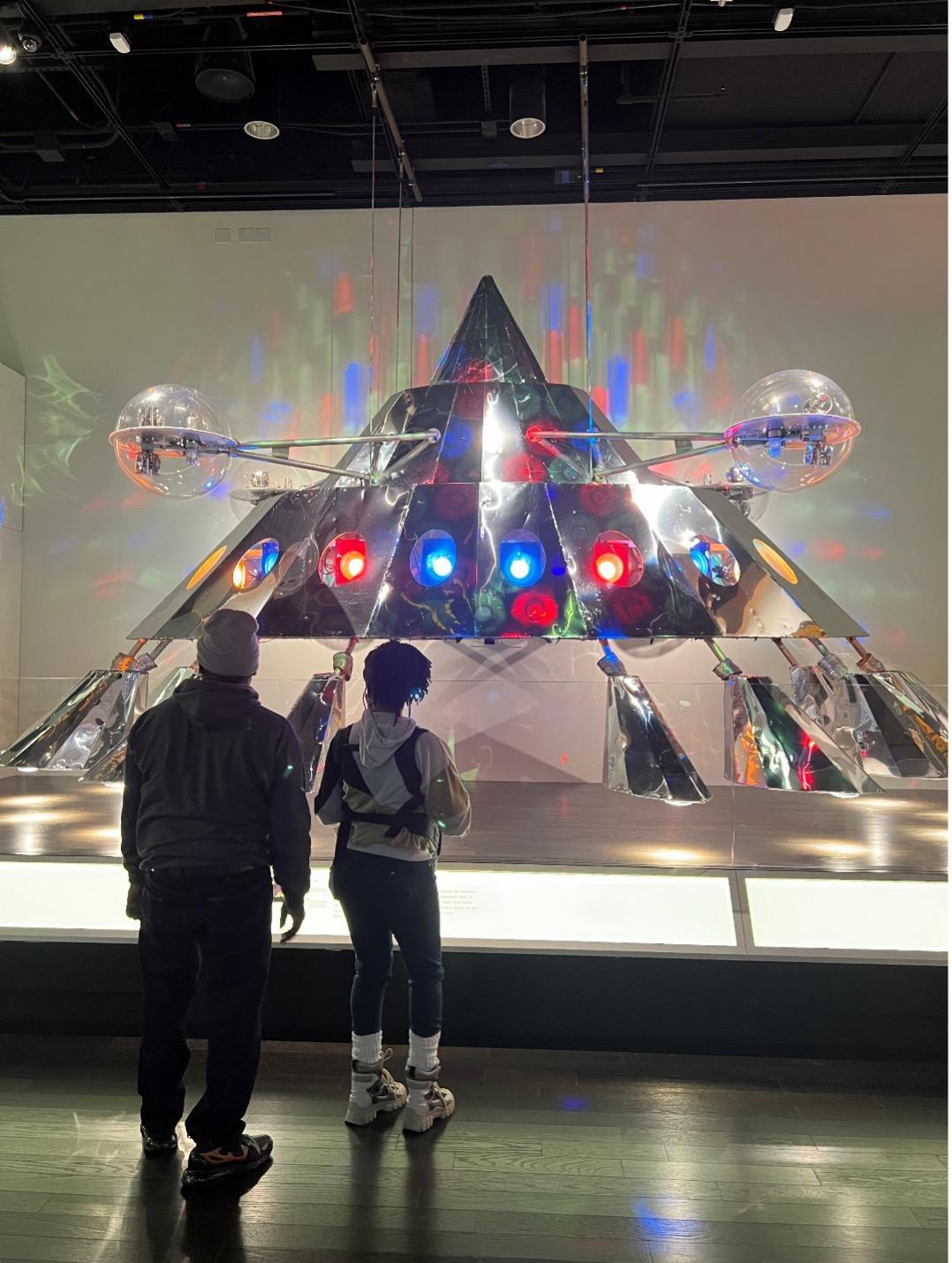 A shiny silver metal triangular spaceship with red and blue lights and eight metal legs is displayed in National Museum of African American History and Culture. Two museum visitors stand in front of the spaceship and read a description panel.
