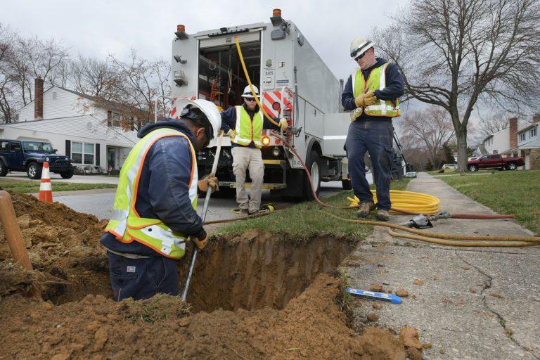 Utility workers watch as on measures in a dirt hole next to a sidewalk.