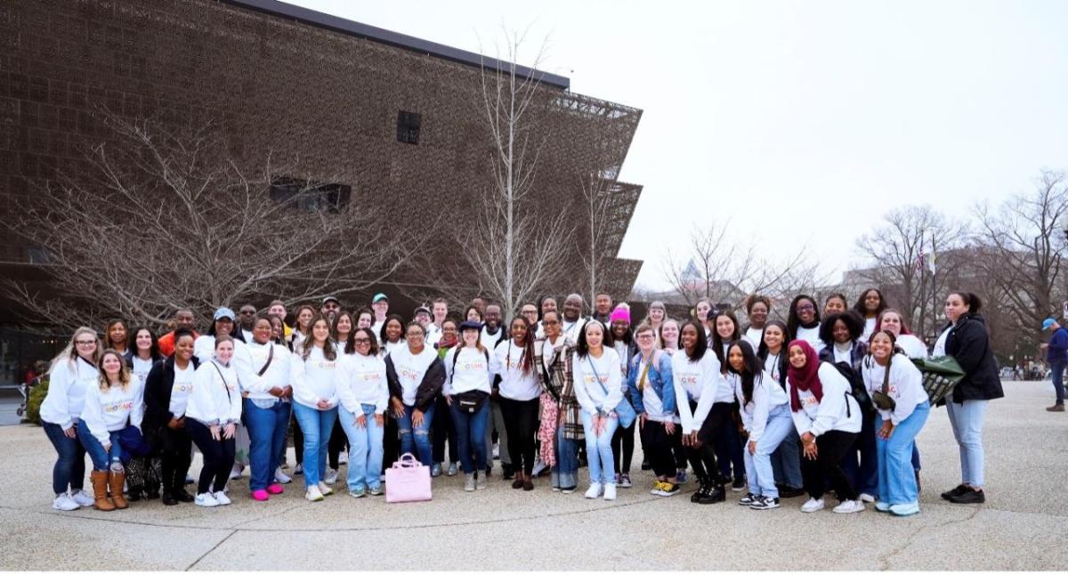Over 50 Bath & Body Works associates are smiling pictured together in matching shirts in front of the National Museum of African American History and Culture. The museum building is in the background.