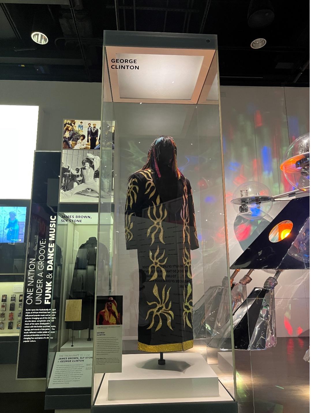A six foot tall museum display case shows a mannequin wearing George Clinton’s black rainbow wig with rainbow accents. The mannequin is also wearing a floor length black robe with golden sunflowers and Swarovski crystals.