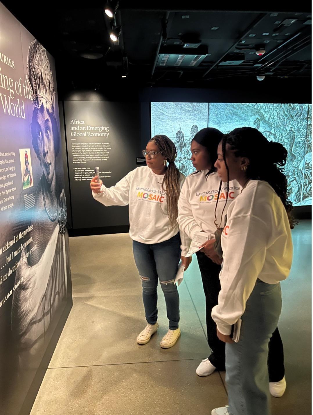 Three women stand in front of museum panels reading history content. One takes a photo with a cell phone as two look on.