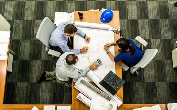 Overhead view of three people looking at blueprints on a shared desk.