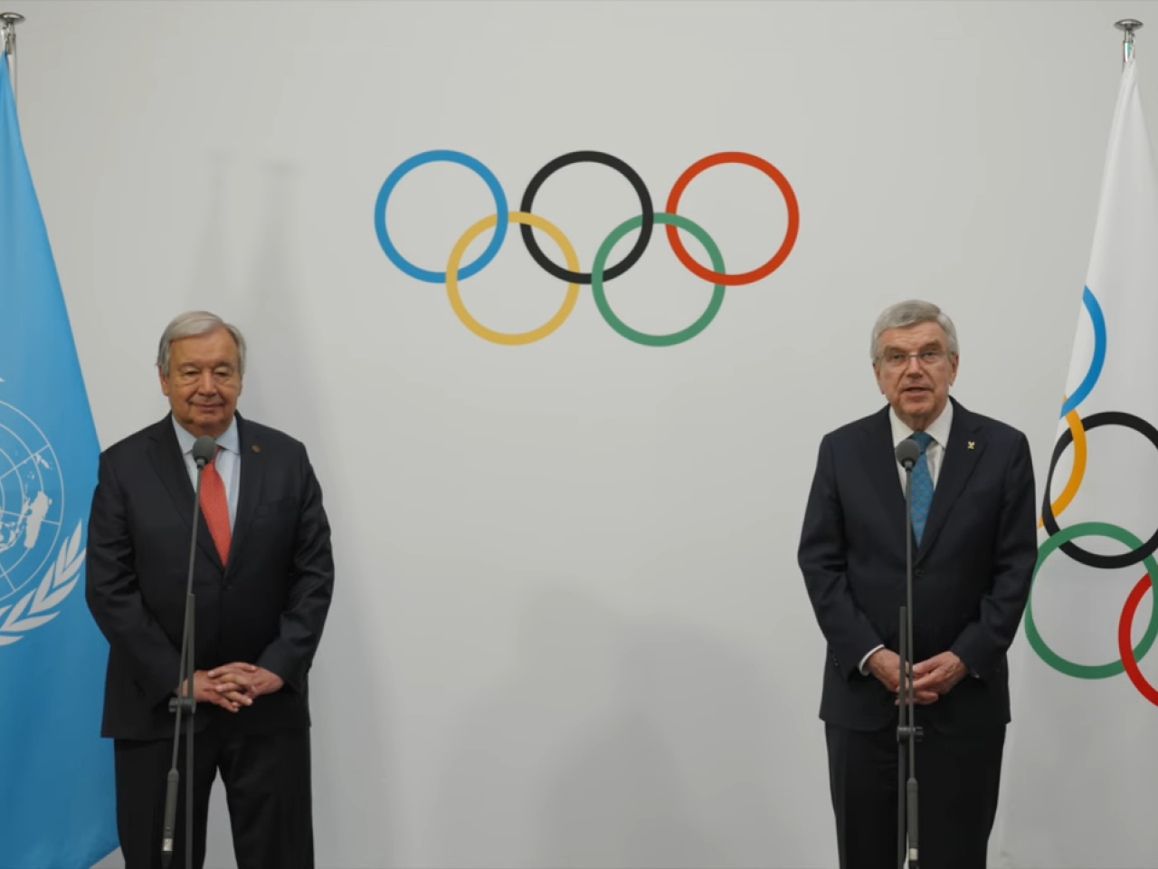 Two people stood in front of an Olympics logo backdrop, talking into a standing microphone 