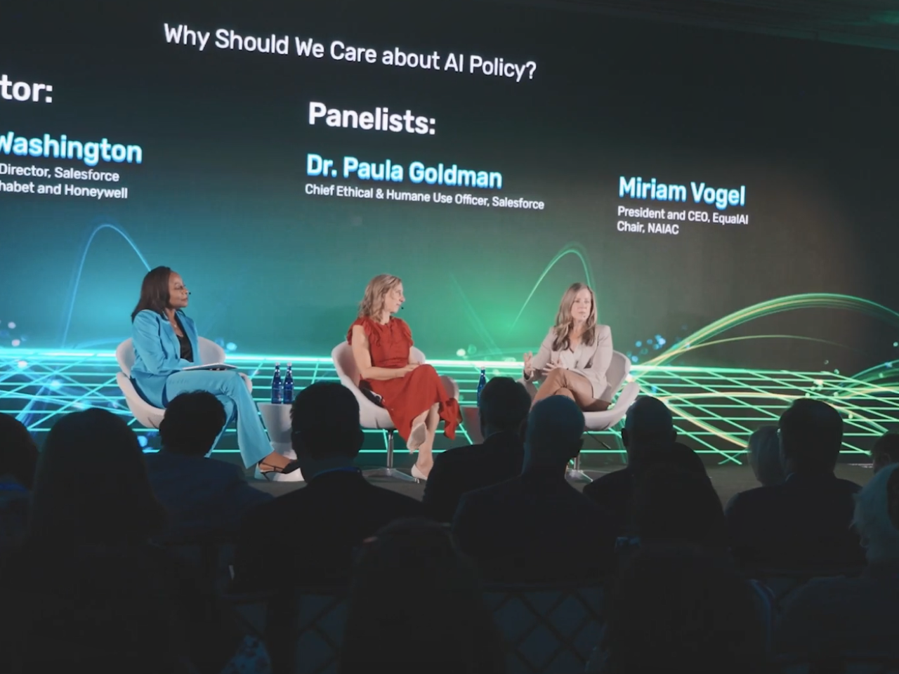 Three seated speakers on a stage in front of a seated audience. Names, titles and "Why should we care about AI policy?" on a screen behind them.