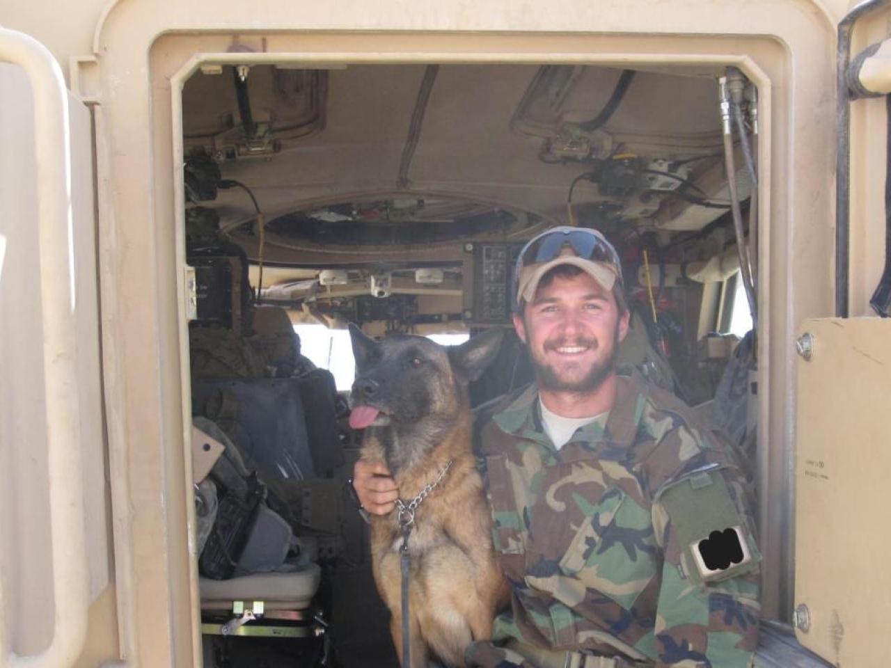 A smiling person in camoflage, sitting in an open vehicle door with a dog