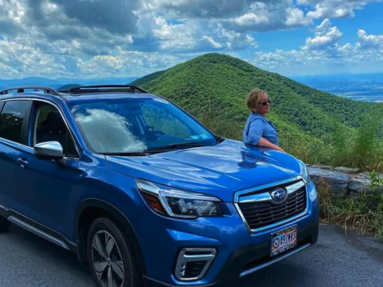 A person leaning against a blue Subaru car, looking out at a long range view