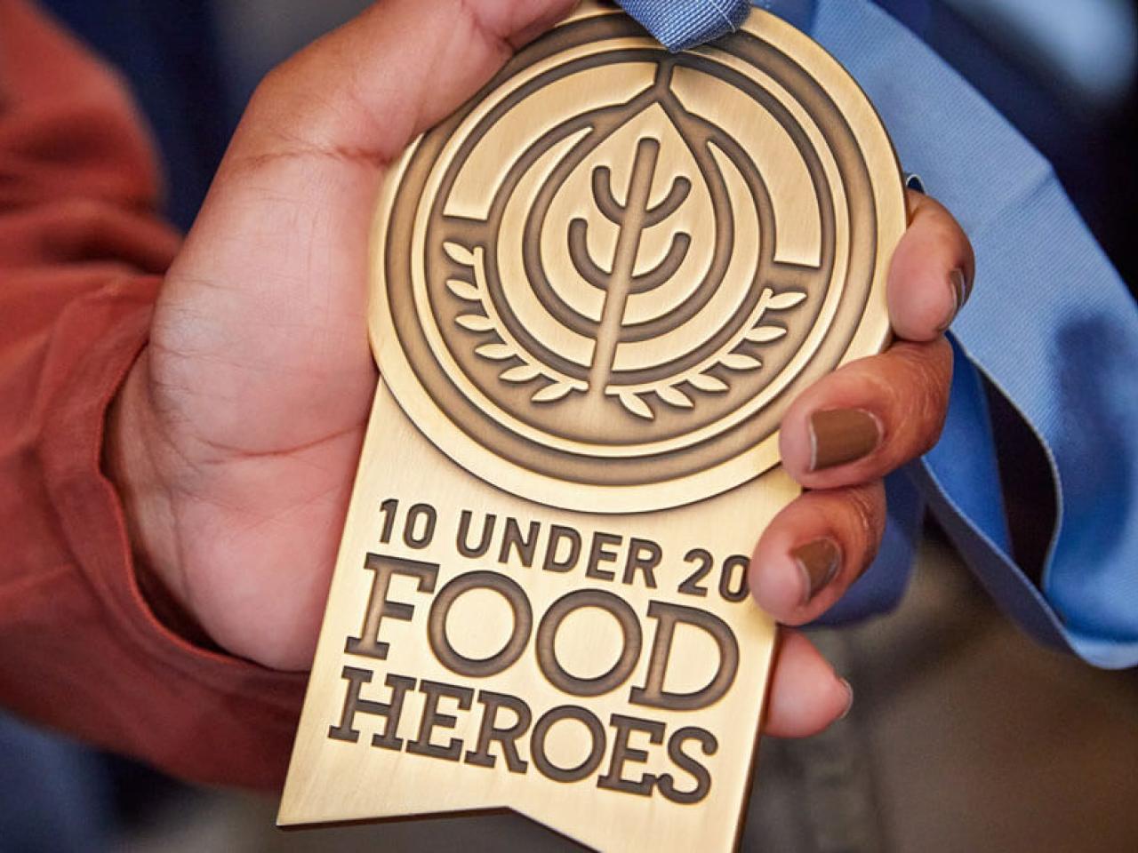 A hand holding a medal "10 Under 20 Food Heroes".