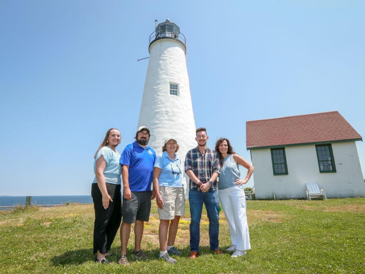 A group of five people pose in front of a light house on an island.