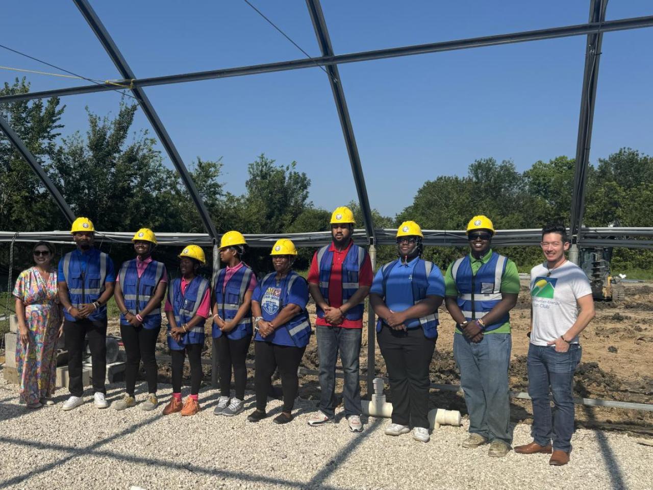 Group of people in greenhouse structure, wearing blue construction vests and yellow hard hats