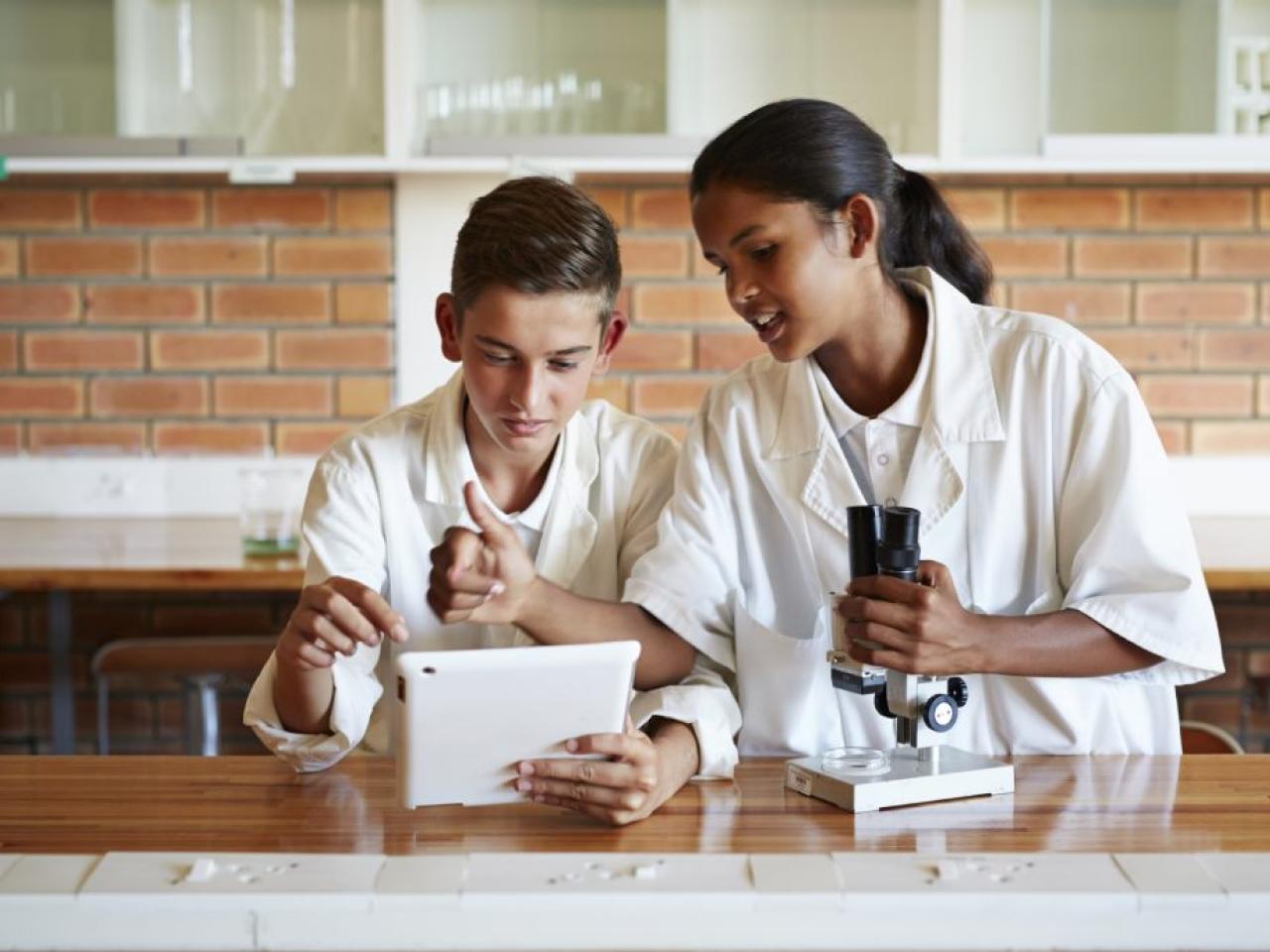 Two young students in a lab looking at a tablet device while another holds a microscope
