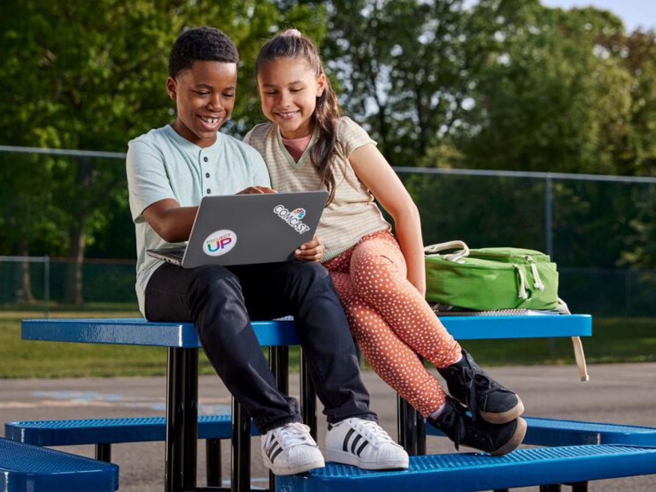 Two smiling children looking at an open laptop while sitting on an outdoor table.