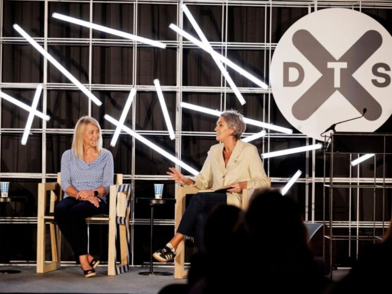 Two people seated on a stage. DTS logo displayed behind them.