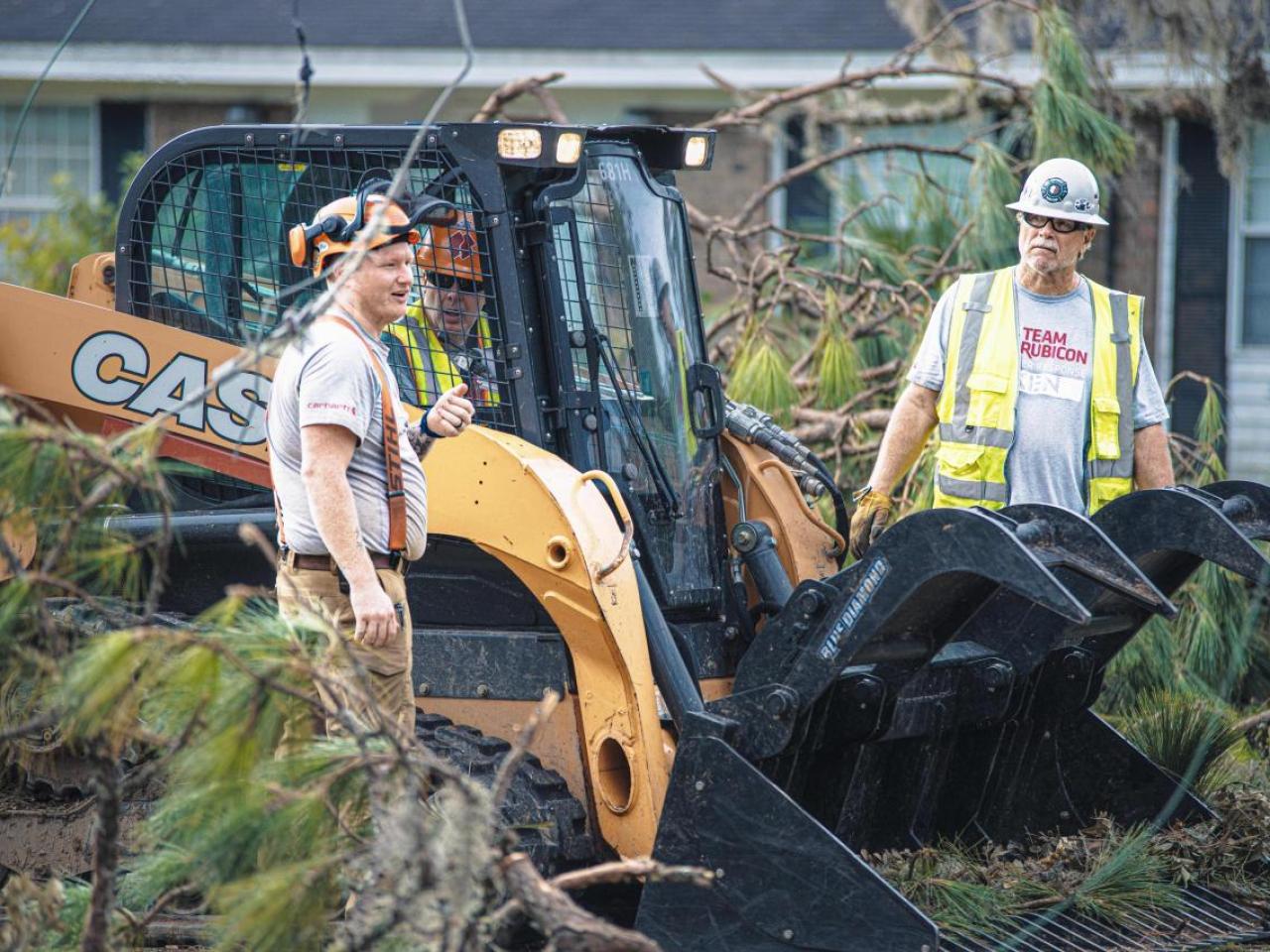 People standing next to a scooping types construction vehicle. Debris and branches surrounding.