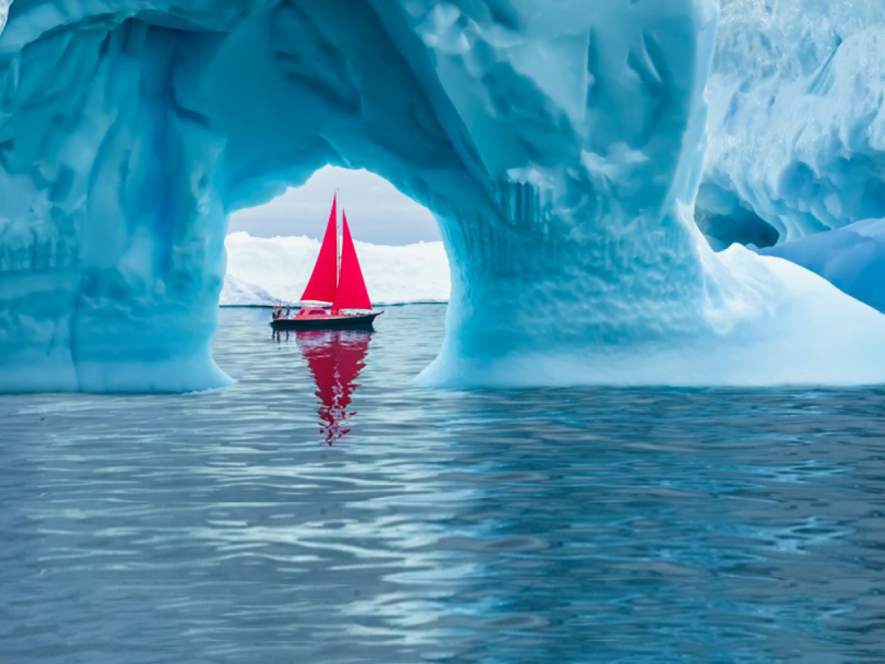 A boat with red sails framed in a small opening in an iceberg on a body of water.