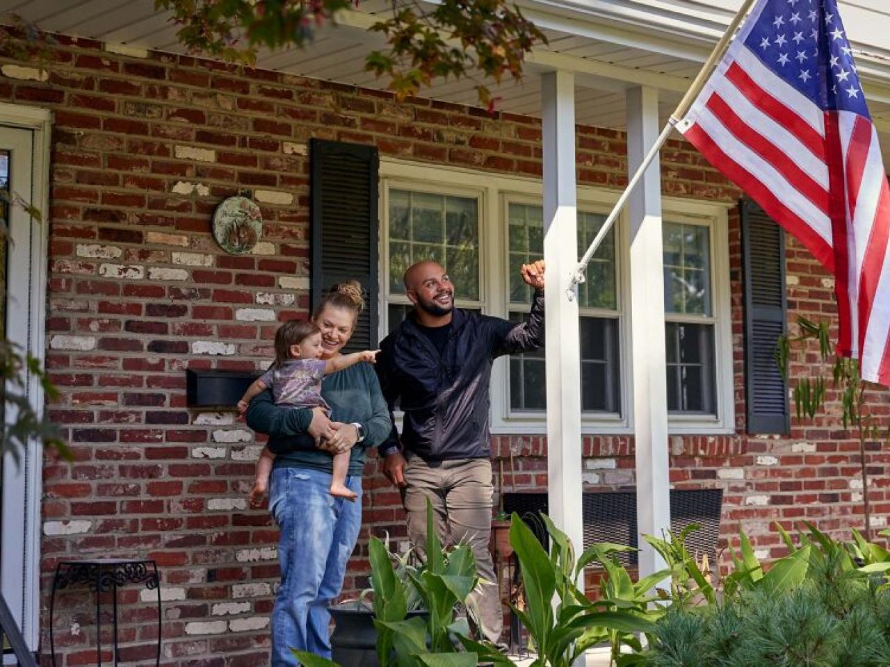 Two adults and a small child standing on a porch. An american flag in a post on the side pillar.