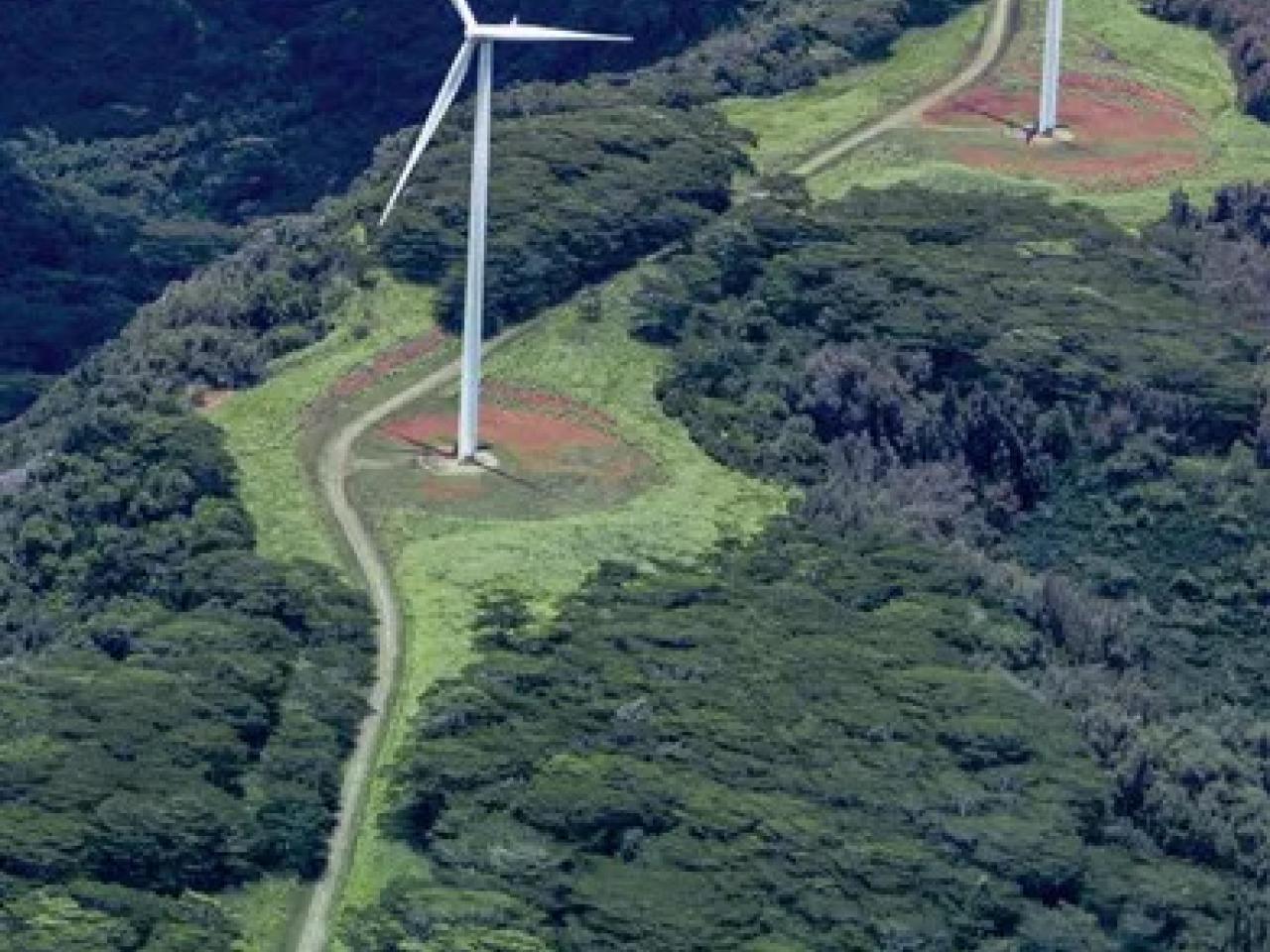 Aerial view of two wind turbines in a mountainous, tree-dense area.