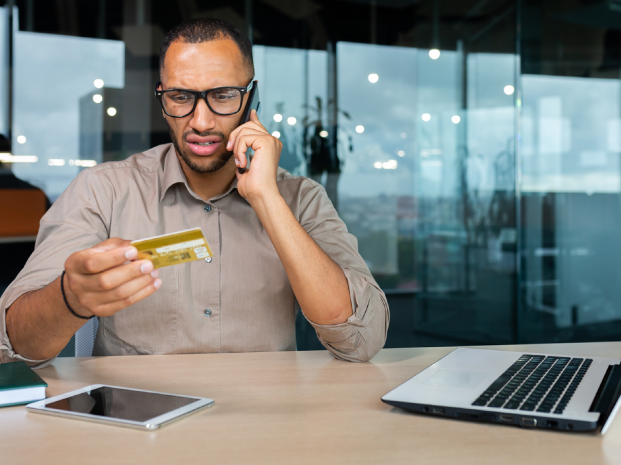 A person talking on a cell phone, looking concerned, holding up a credit card. A laptop computer open next to them on a desk.