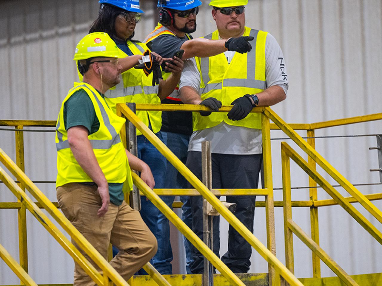 Four employees in hard hats and high-vis vests standing on a platform. One pointing to the distance.