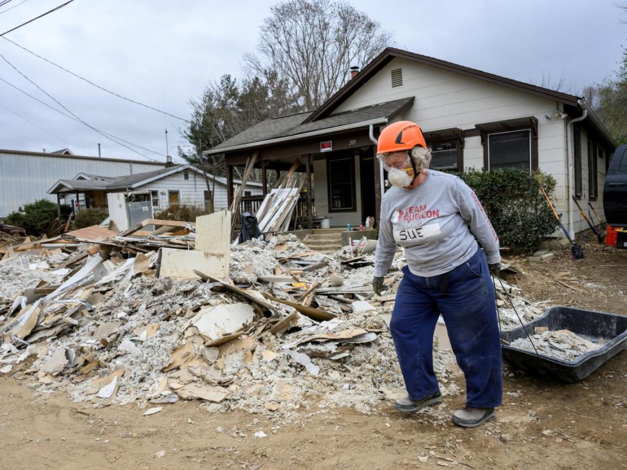 A volunteer in hard hat and respirator pulling a black sled full of rubble. A pile of rubble in front of the house behind them.