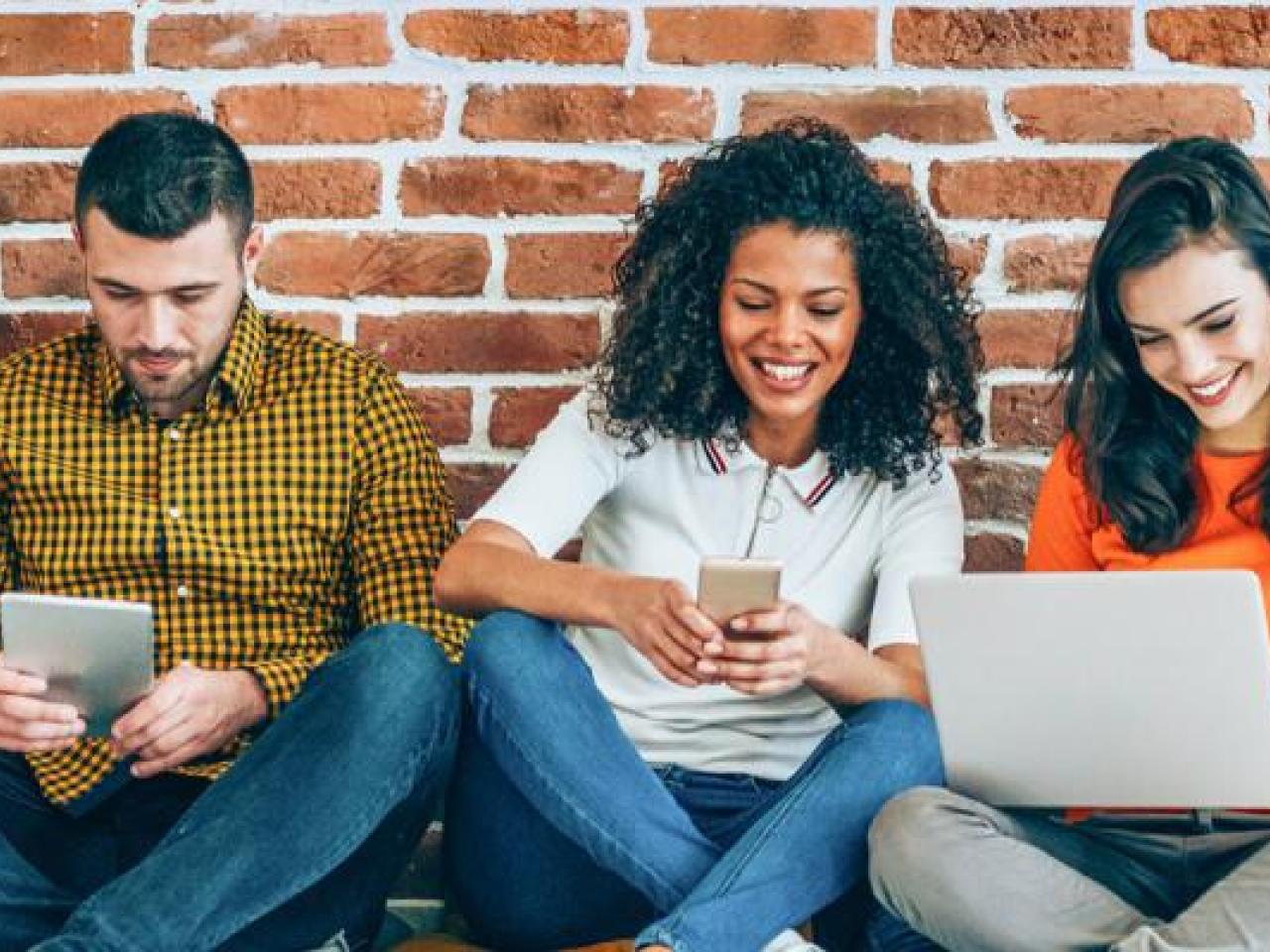 A group seated against a brick wall. Some smiling and looking at an open laptop or other devices.