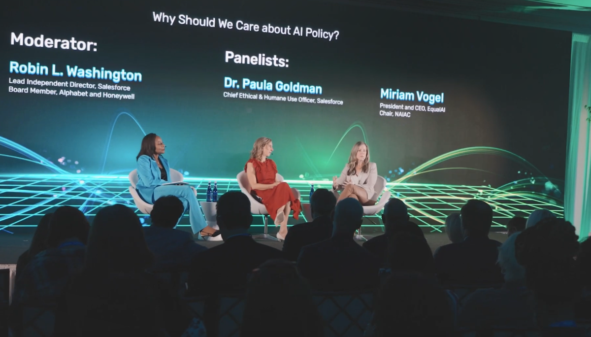 Three seated speakers on a stage in front of a seated audience. Names, titles and "Why should we care about AI policy?" on a screen behind them.