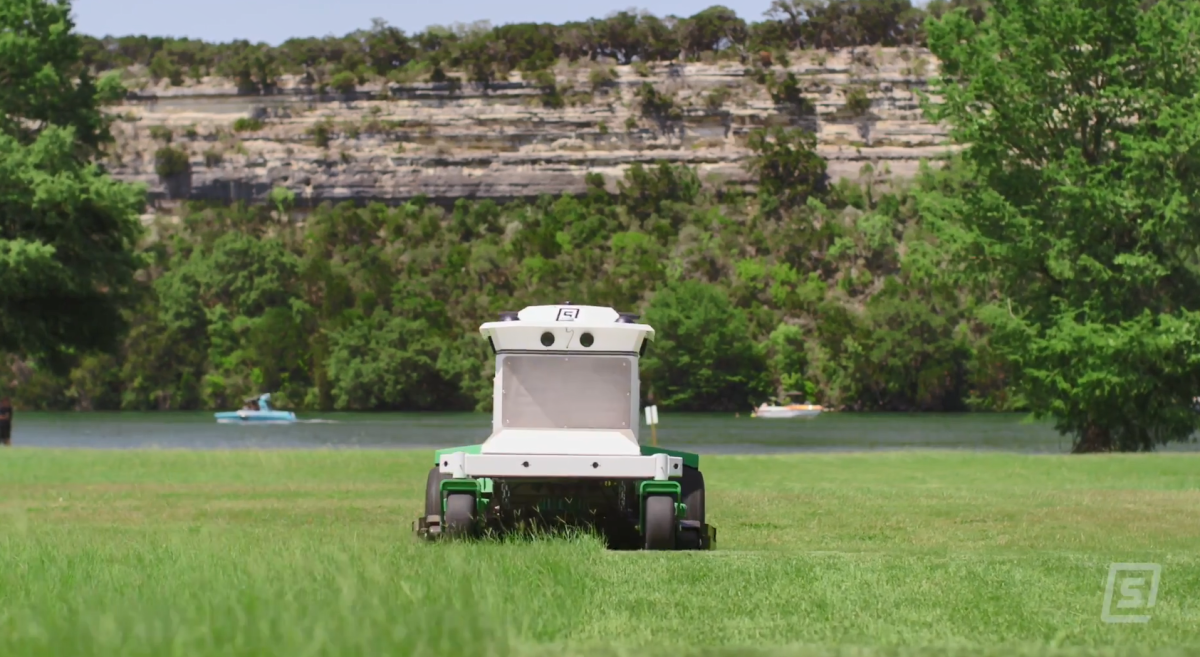 The Scythe mower in action, cutting a large grassy area. Boaters in a body of water behind it and a tall rocky ledge in the background.