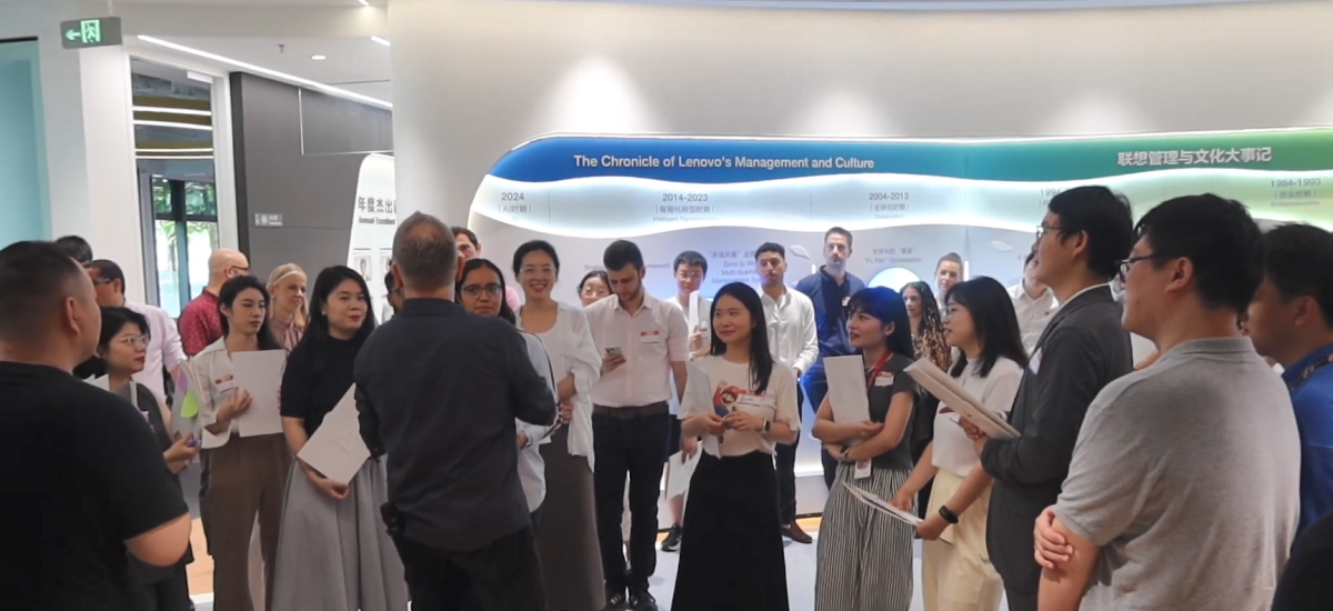 A group gathered around one speaking in an open area with displays on the wall.