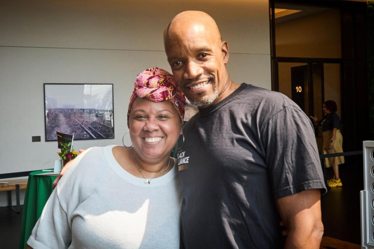A smiling Black woman wearing a white shirt and a colorful scarf top knotted on her forehead and a smiling Black man wearing a black t-shirt