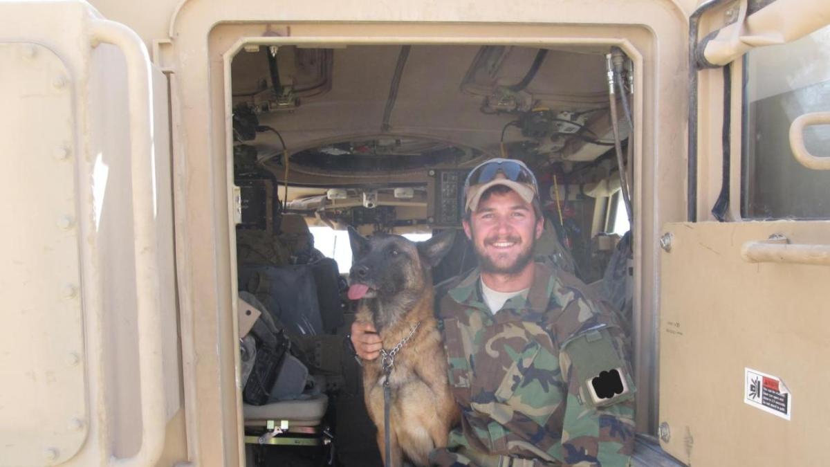 A smiling person in camoflage, sitting in an open vehicle door with a dog