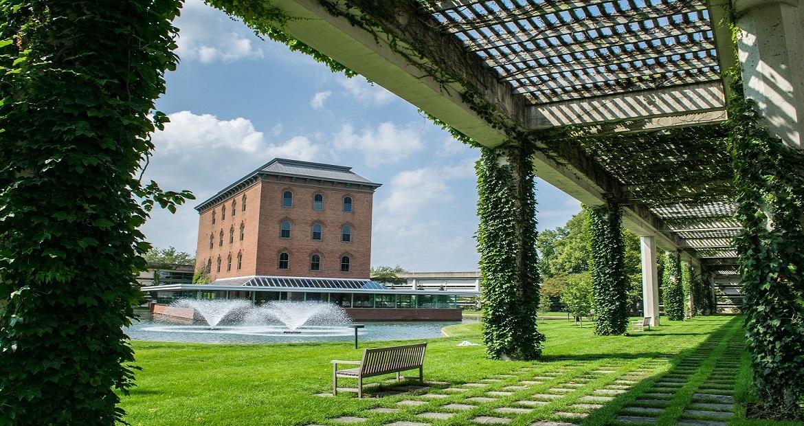 From under a plant-covered pergola, A rectangular brick building behind a water feature.