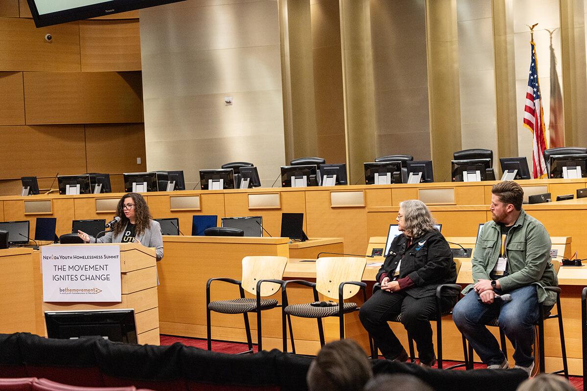 Two people in chairs listening to a speaker behind a podium