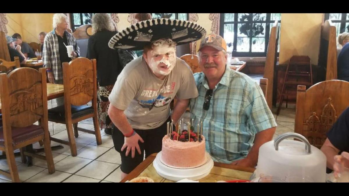 A person in a large sombrero with cream on their face, posing behind a cake with another person sitting at a table