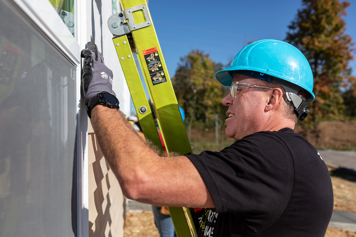 Someone in a blue hard hat next to a ladder working on a house