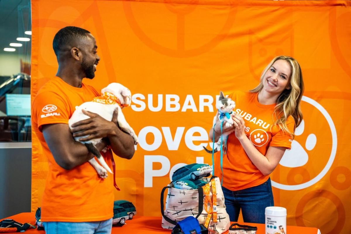 Two people holding a puppy and a kitten, stood in front of an orange Subaru Loves Pets backdrop 