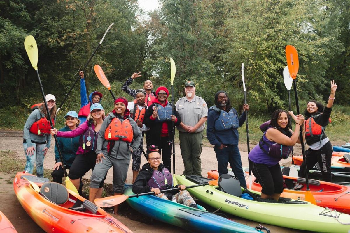 People on shore standing next to colorful kayaks and holding up paddles