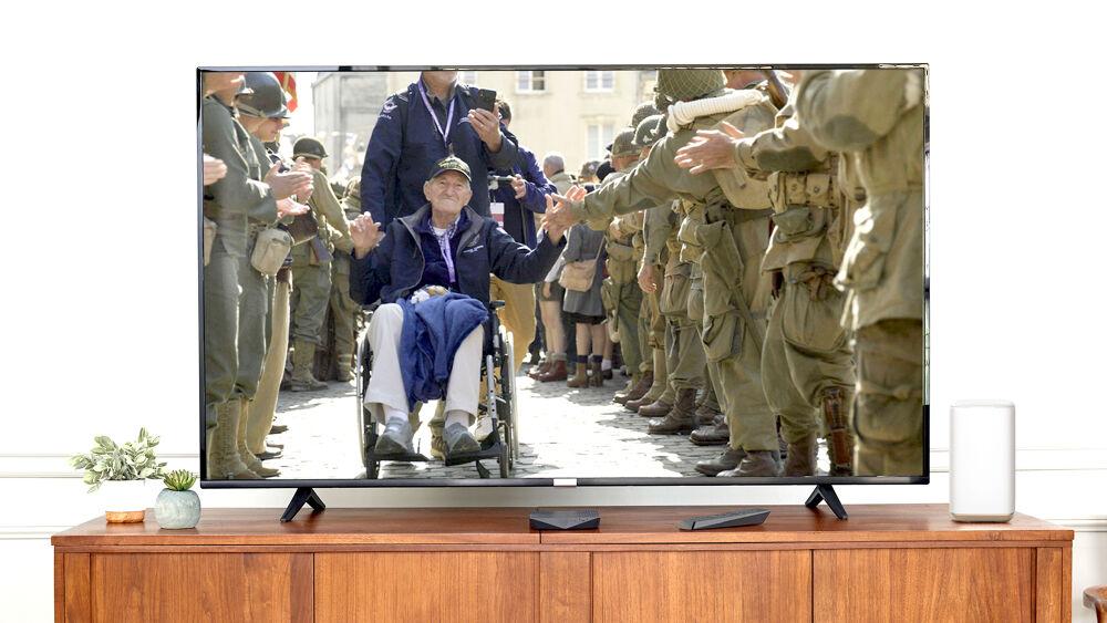 A TV screen on a desk showing an image of a person in a wheelchair being pushed by another as they wave to standing lines of others in military uniform.