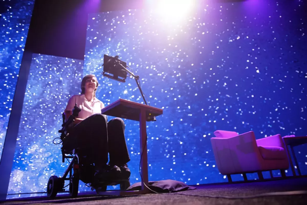 Erin on stage, a digital device in front of them and a colorful starry sky displayed behind.