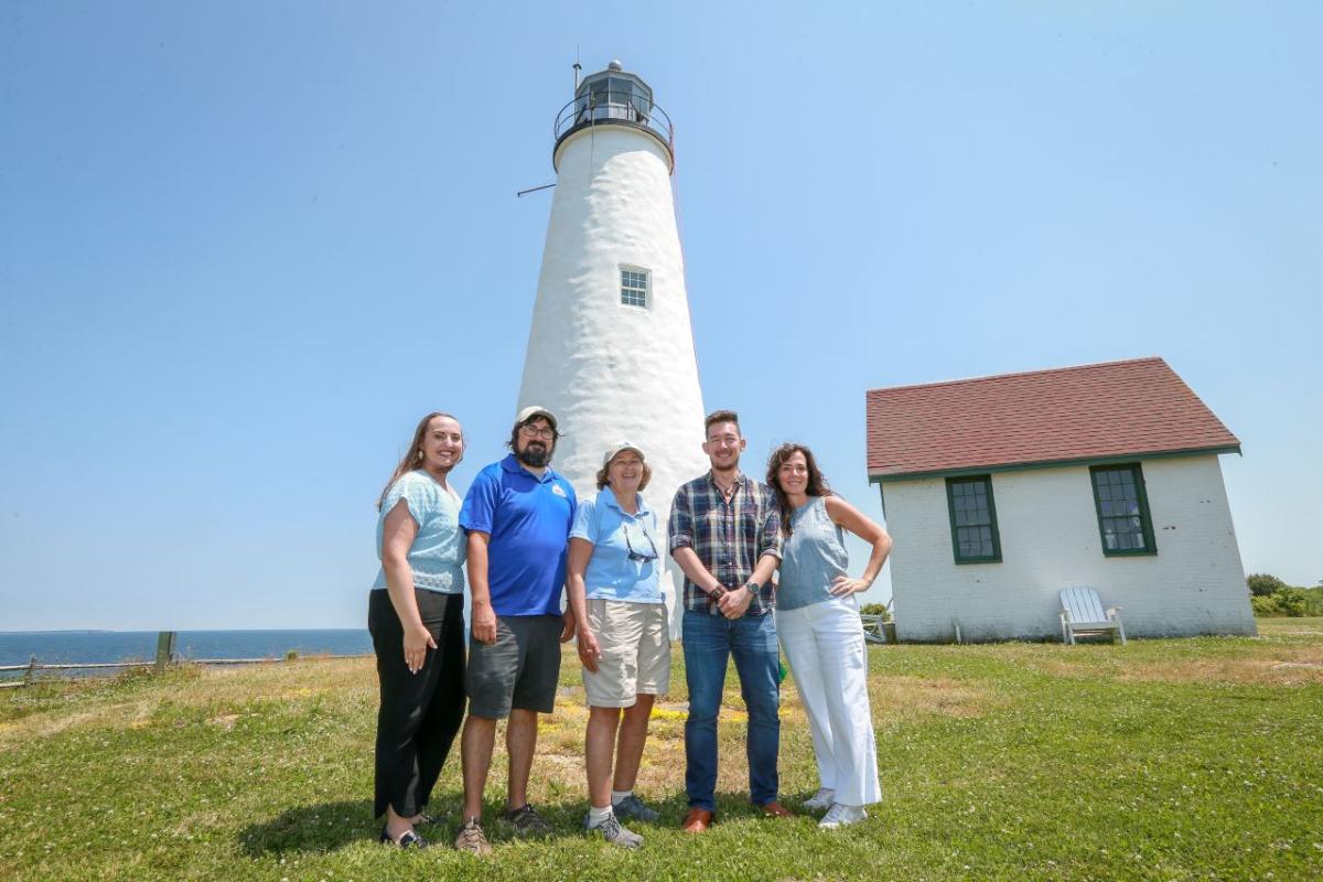 A group of five people pose in front of a light house on an island.