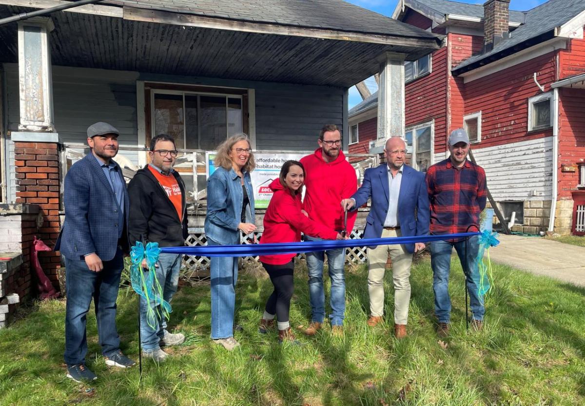 A group posed cutting a blue ribbon in front of a house.