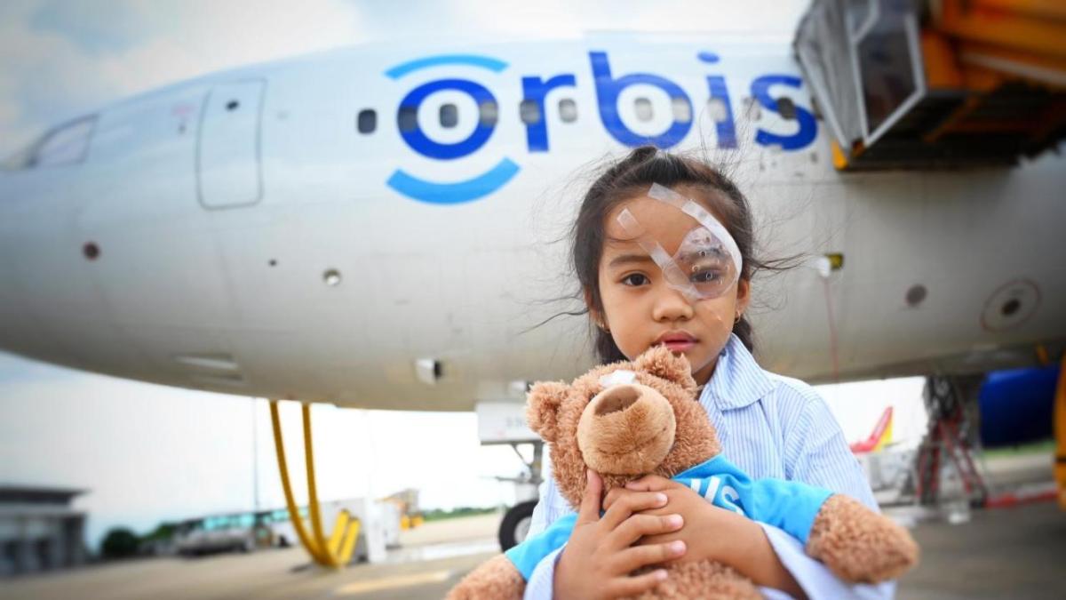 Han wearing an eye patch, holding a teddy bear in front of a plane with Orbis on the side.