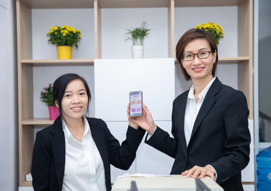 Two people holding a phone up. Shelves behind them each with flowers in them.