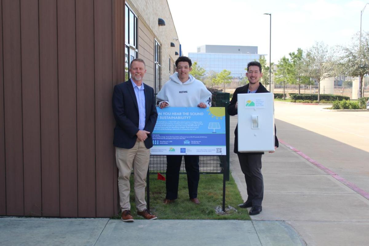 Three men standing around blue sign smiling, with one of them holding an oversized prop light switch