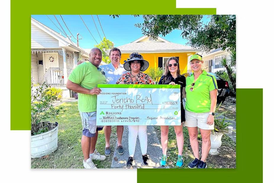 Five people holding a large check in front of a yellow house.