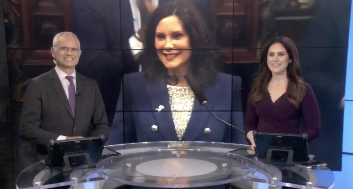 Two news hosts sat at as desk with a screen behind them