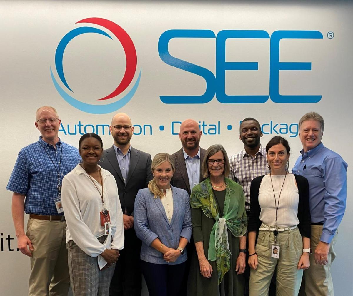 A group of people stand in front of the SEE company logo facing the camera.