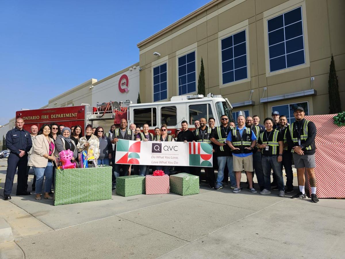 A mix of team members from Qurate Retail Group are standing outside a QVC facility in front of a firetruck along with fire fighters. A few people are holding up a banner which reads "QVC Do What You Love, Love What You Do". They are also surrounded by wrapped gift boxes and holding children's toys as part of a holiday toy drive event.  